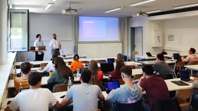 A lecture hall, two men at a computer in the front, a presentation on the screen next to them, a large number of students opposite.