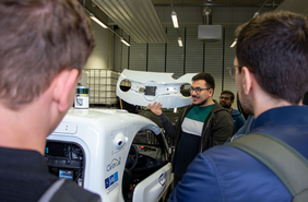 Ömer Dönmez shows a group something in the lab in front of an autonomous vehicle