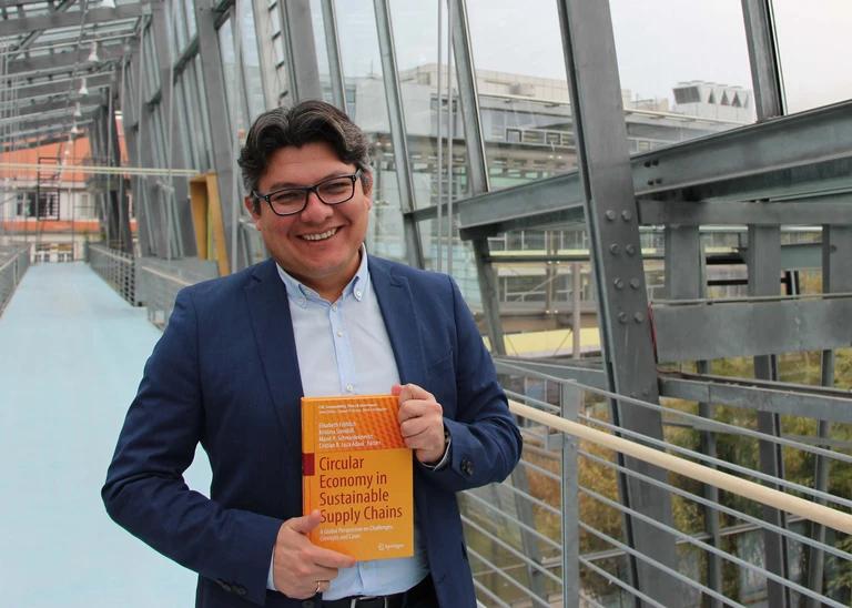 A man in a suit shows an orange book, with a glass THI building in the background.