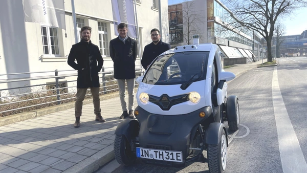 Ein kleines Auto steht an der Straße, im Hintergrund stehen drei Männer