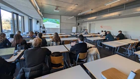 A lecture hall from behind, you can see the speaker with a PowerPoint presentation in front.