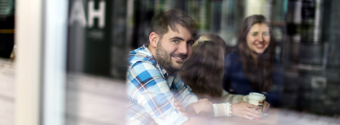 Three students sit behind a window front, one of them smiles into the camera