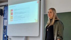 : A speaking woman on the right edge of the picture, to her left a presentation on the blackboard.