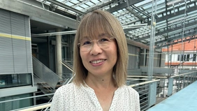 A Japanese woman smiles into the camera, in the background the modern glass building of the THI.